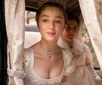 Young woman in elegant gown and tiara looks out a carriage window with another woman in the background.