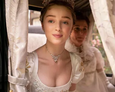 Young woman in elegant gown and tiara looks out a carriage window with another woman in the background.