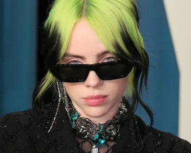 Woman with black sunglasses and a green-black hairstyle wearing a sparkling black outfit and jewelry.