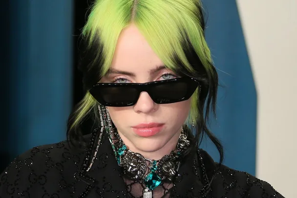 Woman with black sunglasses and a green-black hairstyle wearing a sparkling black outfit and jewelry.