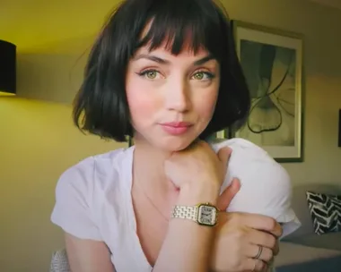 A person with a short black haircut and bangs, wearing a white shirt and watch, sitting indoors.