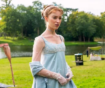 A woman in a Regency-era blue dress stands by a lake with trees in the background.