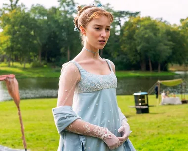 A woman in a Regency-era blue dress stands by a lake with trees in the background.