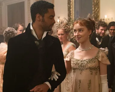 A couple in Regency-era attire dances at a formal ball, with decorative flowers and mirrors in the background.