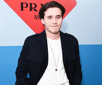 Smiling man in black jacket and white shirt, wearing a pendant necklace, standing against blue and red background.