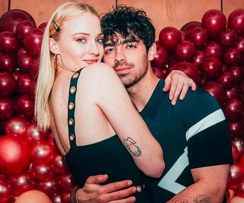 A couple embraces in front of a red balloon backdrop; woman's shoulder tattoo visible on the left.