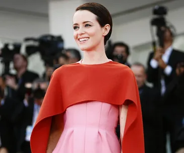 "Claire Foy smiling in a pink dress with red cape at a public event, surrounded by photographers."