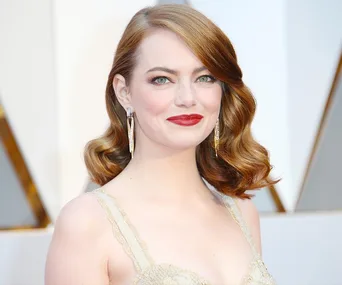 Smiling woman with wavy auburn hair, wearing a gold dress and statement earrings, against a blurred light background.