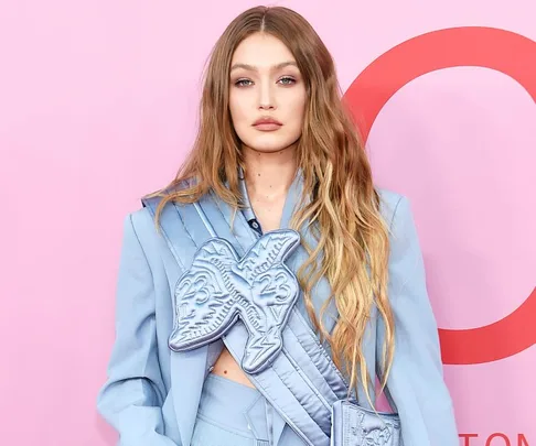 Model in a light blue suit with long wavy hair, standing against a pink backdrop.