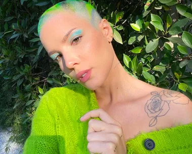 A person with bright green hair and eye makeup poses in a green sweater, showing a rose tattoo on their shoulder.