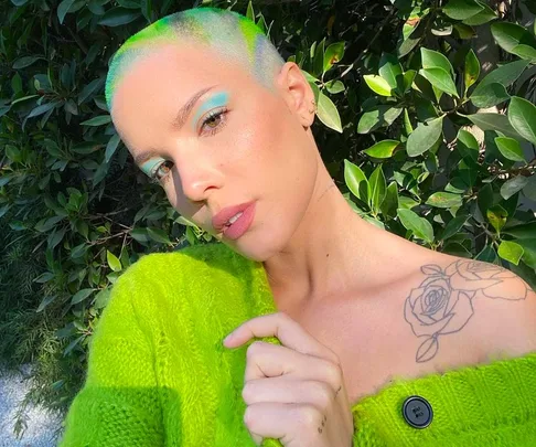 A person with bright green hair and eye makeup poses in a green sweater, showing a rose tattoo on their shoulder.