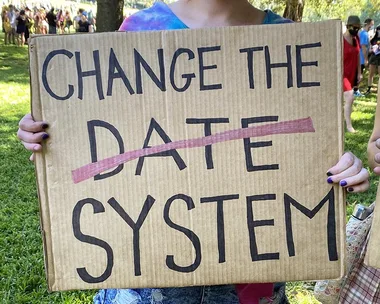 Protester holds a cardboard sign reading "CHANGE THE ~DATE~ SYSTEM" during an outdoor rally.