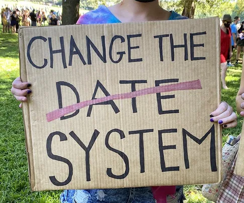 Protester holds a cardboard sign reading "CHANGE THE ~DATE~ SYSTEM" during an outdoor rally.