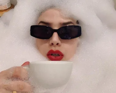 Person with red lipstick wearing sunglasses holds a cup, surrounded by bubble bath foam.