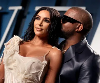 A couple poses together, with the man wearing sunglasses and the woman in a white dress, against a blurred blue background.