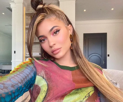 A woman taking a selfie indoors, wearing a colorful top with hoop earrings, styled hair, and makeup.