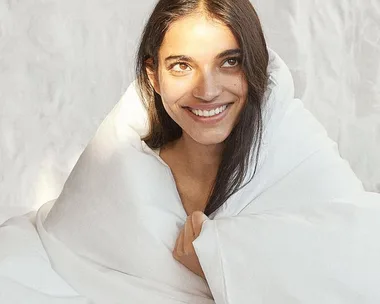 Smiling woman wrapped in a white blanket, looking upwards with bright natural lighting.