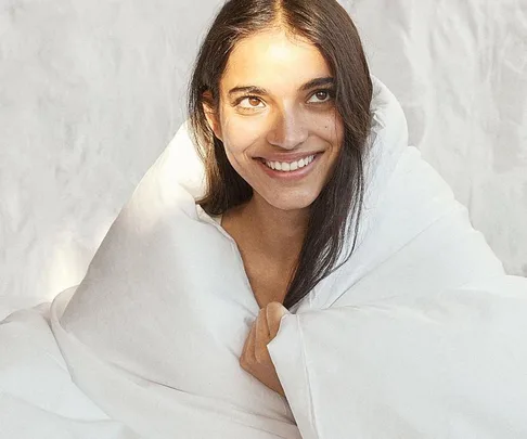 Smiling woman wrapped in a white blanket, looking upwards with bright natural lighting.