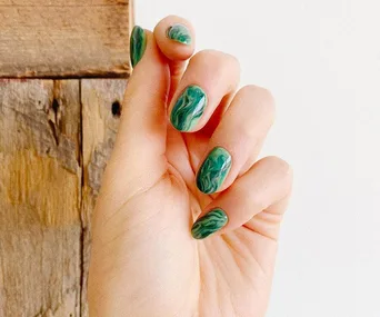 Hand with green marble-style nail art in front of a wooden background.