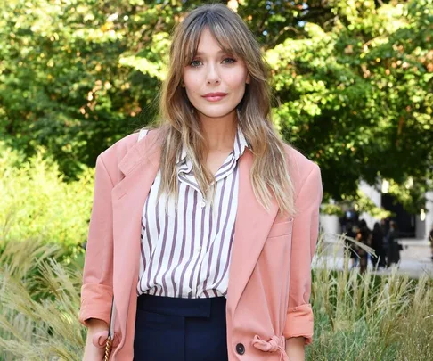 Woman in a pink blazer and striped shirt, standing outdoors with greenery in the background.