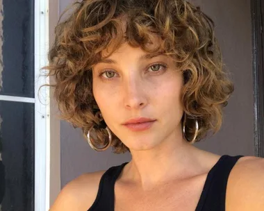 Woman with short, curly hair and hoop earrings, wearing a black top, standing in front of a gray door.