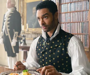 Rege-Jean Page as the Duke of Hastings in Bridgerton, sitting at a dining table in period attire.