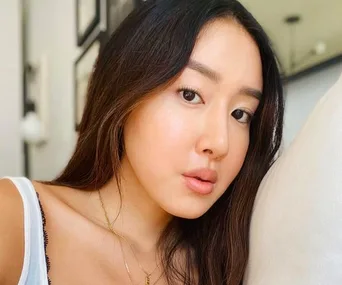 A woman with long dark hair and natural makeup poses indoors, wearing a white top.