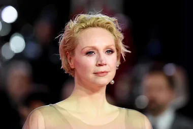 Smiling woman with short blonde hair at a formal event, wearing a sheer dress, with lights blurred in the background.