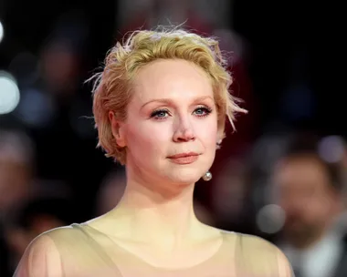 Smiling woman with short blonde hair at a formal event, wearing a sheer dress, with lights blurred in the background.