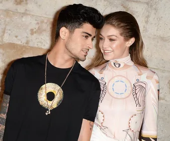 A couple standing close together, smiling at each other, dressed in stylish outfits against a neutral background.