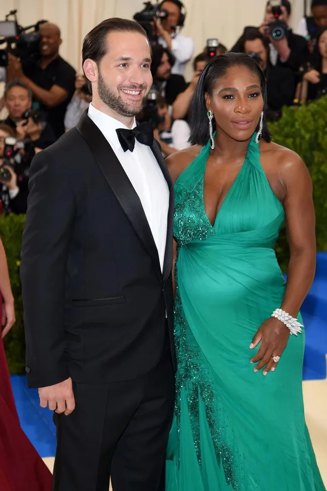 Two people pose together on a red carpet, one in a tuxedo and the other in a green gown, with photographers in the background.