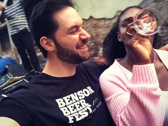Man smiling while woman drinks from a glass, enjoying a casual outdoor setting.