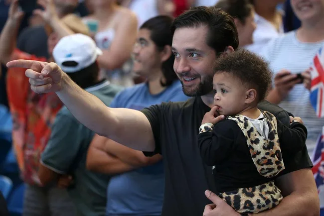 Man holding a child dressed in leopard print, pointing and smiling amidst a crowd at an event.