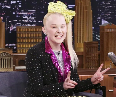 Jojo Siwa on a talk show set, smiling, wearing a studded suit with a large yellow bow and pink sequined top.