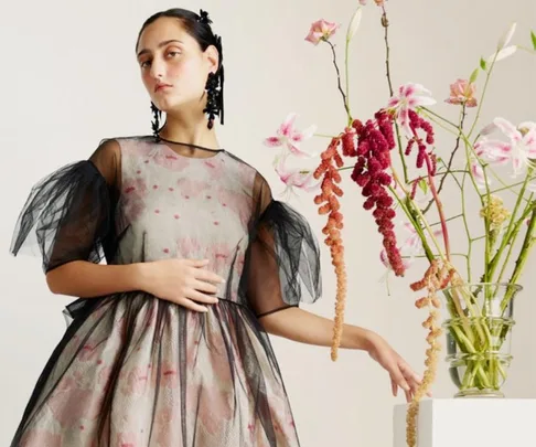 Model in floral Simone Rocha x H&M dress with sheer sleeves, standing by a vase of colorful flowers.