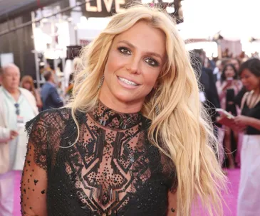 A woman with long blonde hair in a black lace dress smiling at a public event, with people in the background.