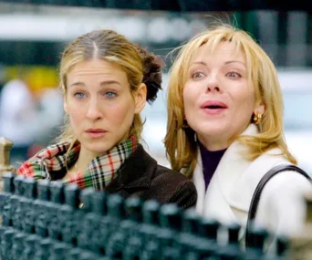 Two women stand outside, one with a scarf and concerned expression, the other smiling widely with short blonde hair.