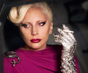 A character from American Horror Story: Hotel, wearing a jeweled glove, holding a cigarette, and looking pensive.