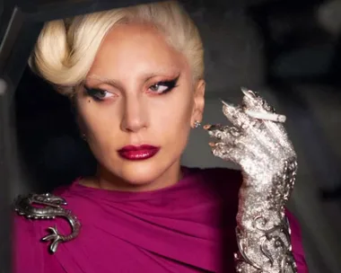 A character from American Horror Story: Hotel, wearing a jeweled glove, holding a cigarette, and looking pensive.