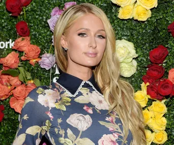 Woman in floral dress stands in front of a wall of colorful roses, looking forward.