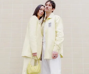 Couple in matching pale yellow outfits against a tiled wall, holding a small yellow handbag.