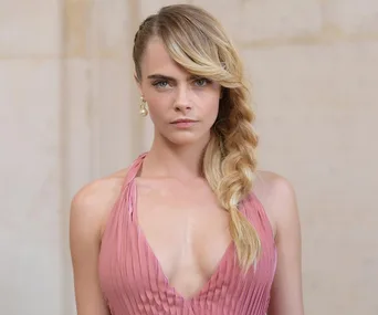 A person wearing a pink dress with a braided hairstyle, looking directly at the camera, against a plain background.