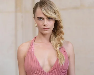 A person wearing a pink dress with a braided hairstyle, looking directly at the camera, against a plain background.