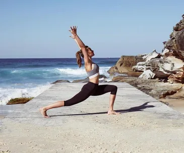 Person doing yoga on a beach, ocean waves in the background, wearing black leggings and gray sports bra.