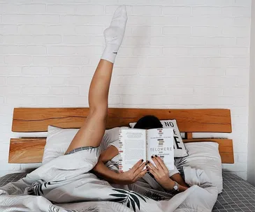 Person lying in bed with one leg up, reading a book titled "Telomere," beside a white brick wall.