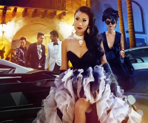 A glamorous group of individuals in formal attire with luxurious cars in the background from "Bling Empire."