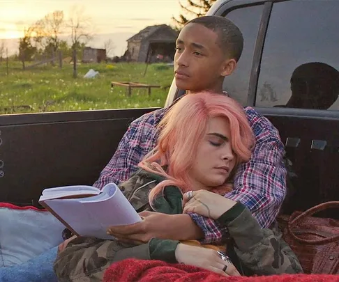 Two people sitting in a truck bed, one with pink hair asleep, holding a script, during sunset in the countryside.