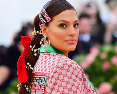 A woman in a patterned Gucci outfit, adorned with pearls and floral hair accessories, attends an event with a floral backdrop.