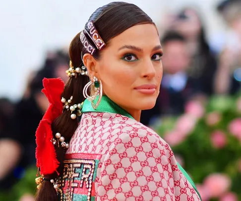 A woman in a patterned Gucci outfit, adorned with pearls and floral hair accessories, attends an event with a floral backdrop.