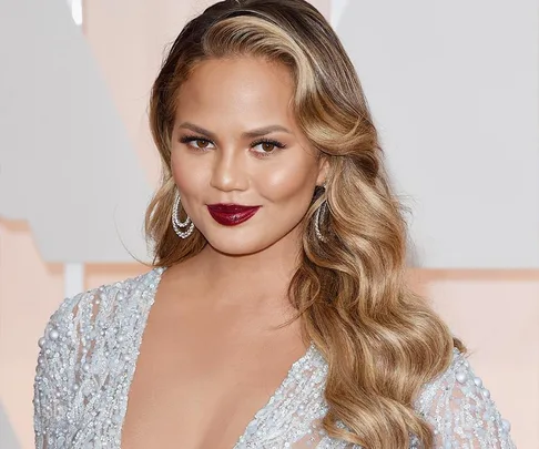 Woman in elegant dress with long wavy hair and bold lipstick, posing on a formal occasion backdrop.